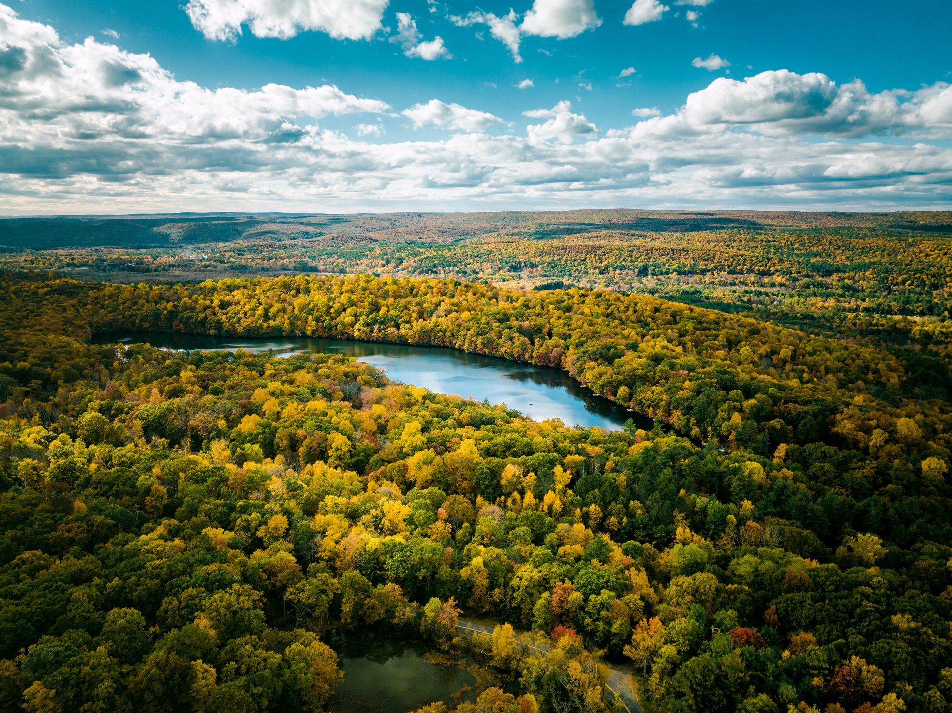 Aerial view of a lake surrounded by a colorful forest in autumn, under a partly cloudy blue sky.