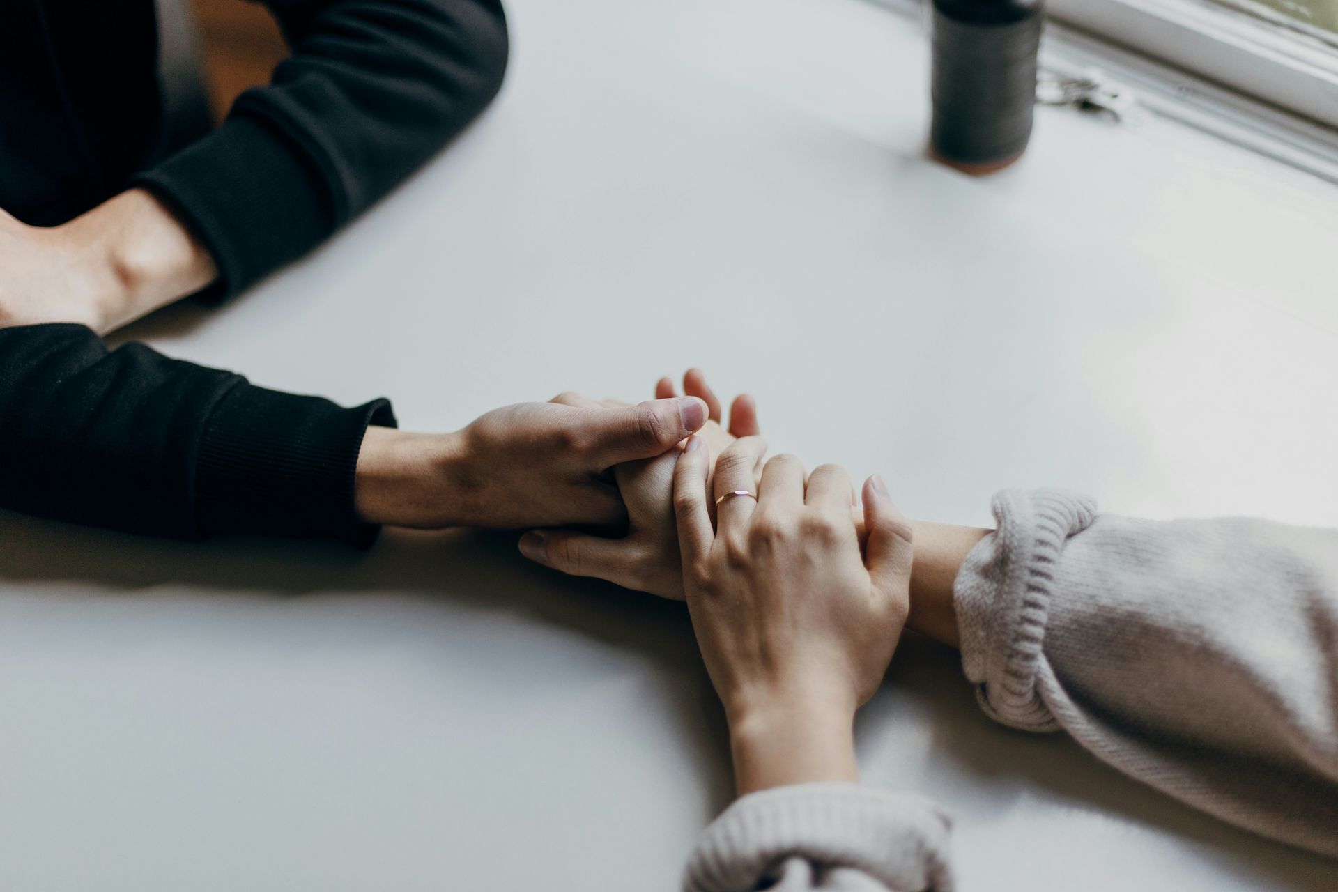 Two people holding hands on a white table. One person wears a black sweater, the other a light sweater.