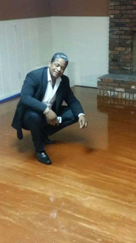 Man in suit crouches on a shiny wooden floor near a brick fireplace.
