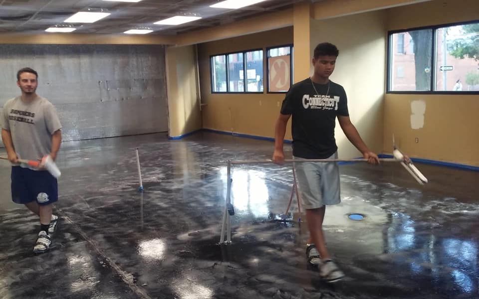 Two people applying epoxy flooring to a room; one using a roller, the other a leveling tool.