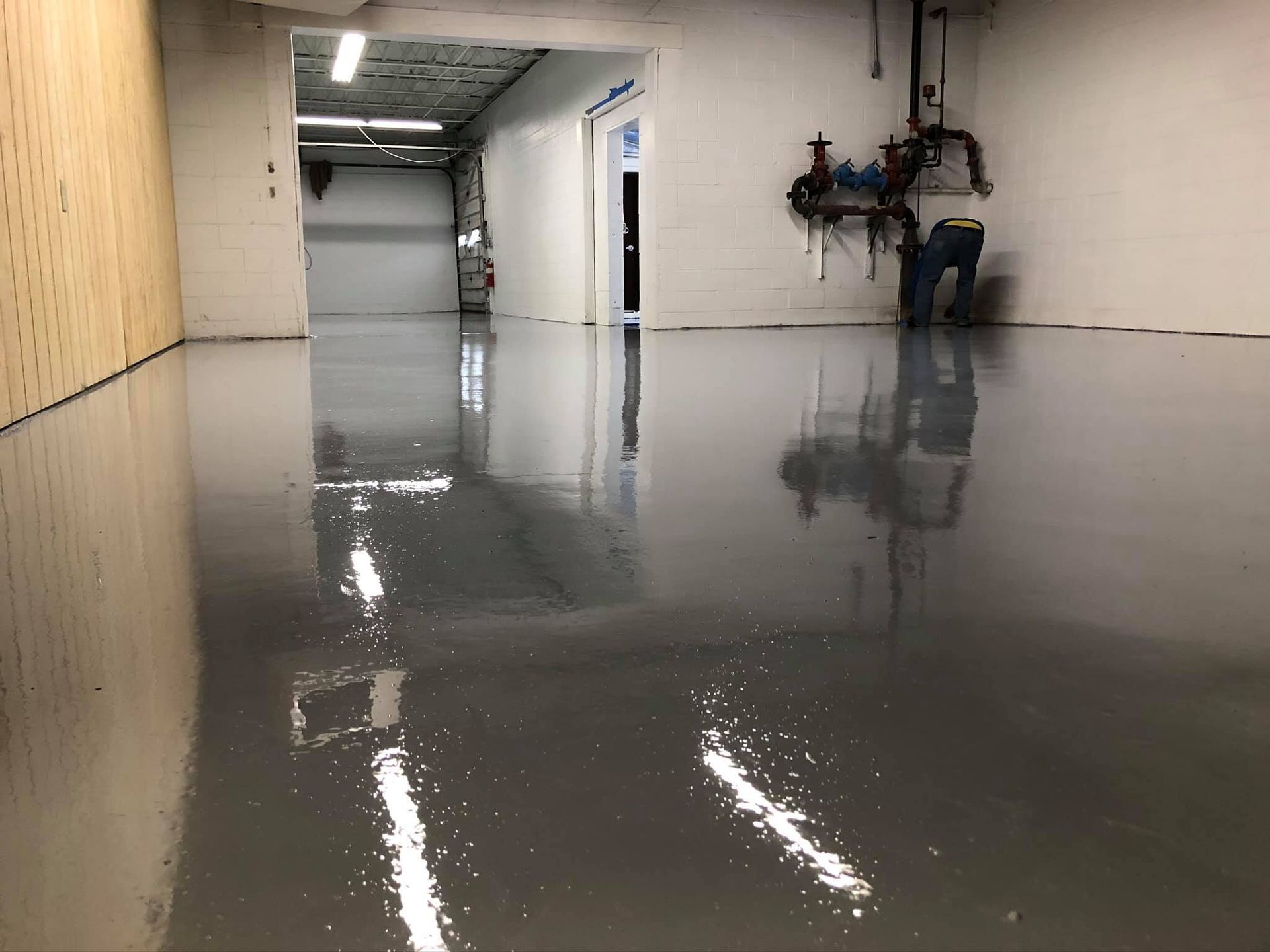 Gray epoxy floor reflects light; a person works near plumbing on a white wall in a utility room.