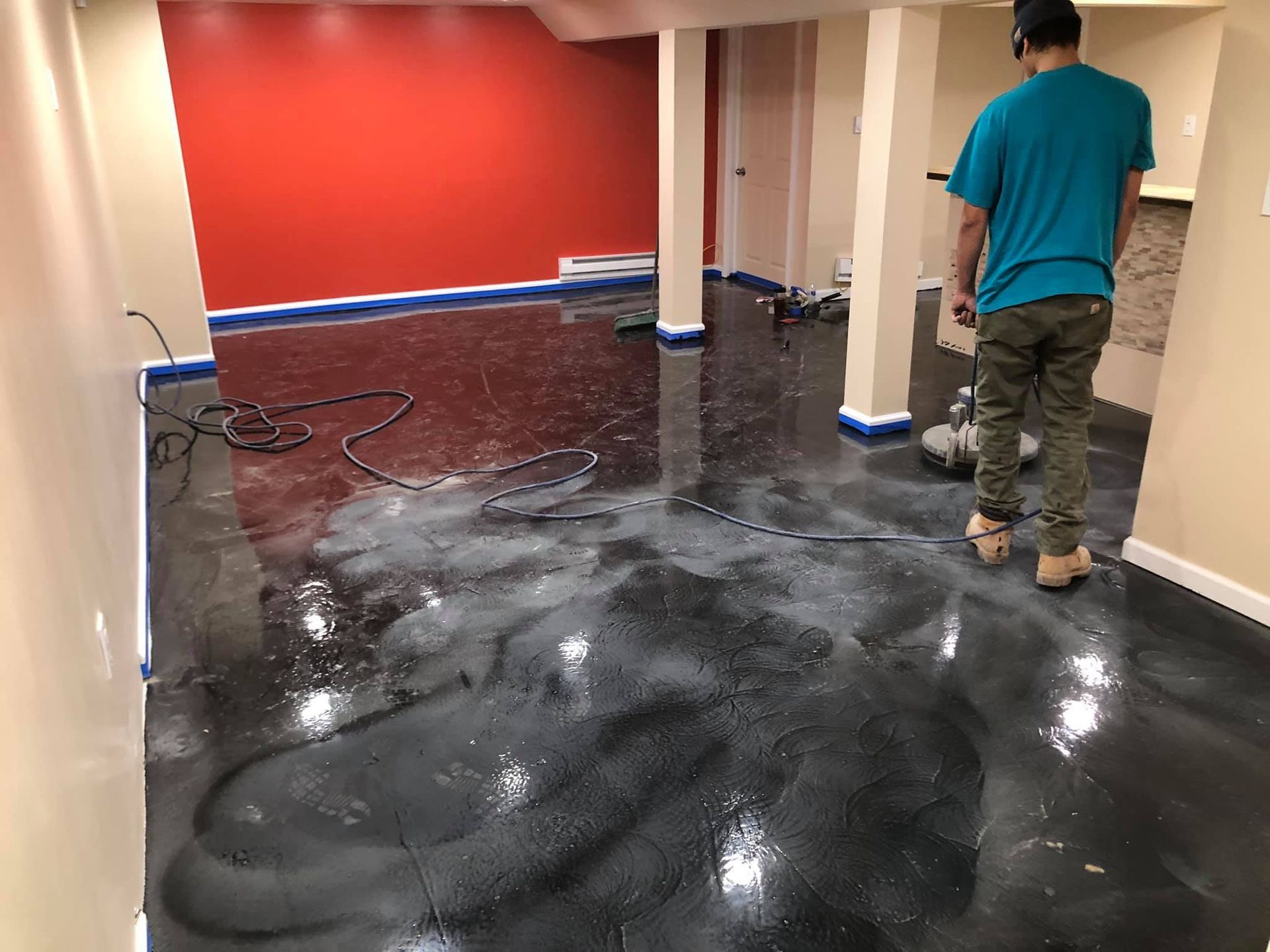 Person refinishing a dark epoxy floor in a basement with equipment; red and beige walls visible.