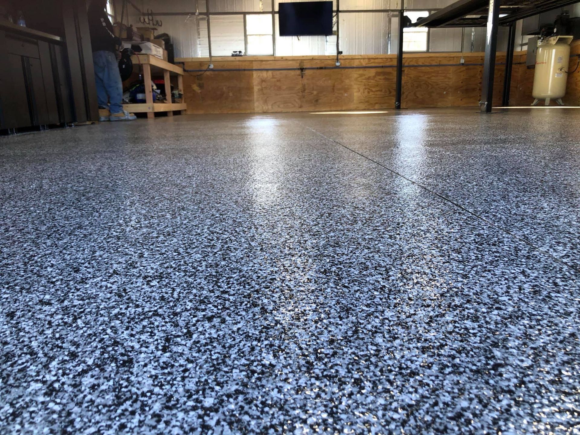 Close-up of a garage floor with a speckled epoxy coating, reflecting the garage's interior and a person in the background.