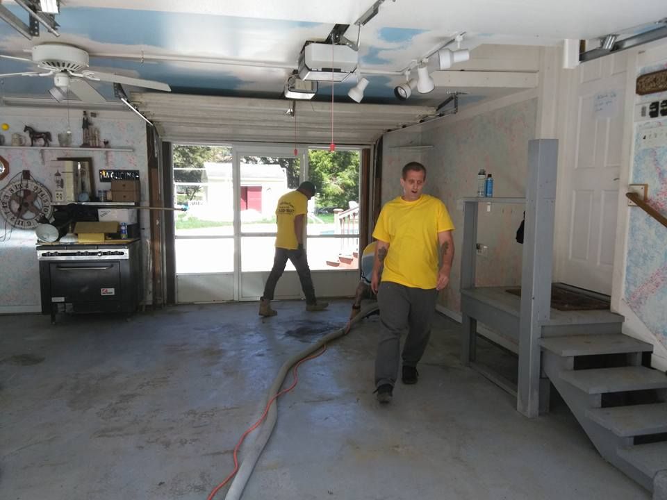 Two men in yellow shirts working in a garage, one with a hose.