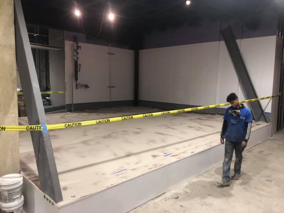 Construction site with caution tape. A worker walks by a concrete structure with metal beams. Walls are white and grey.