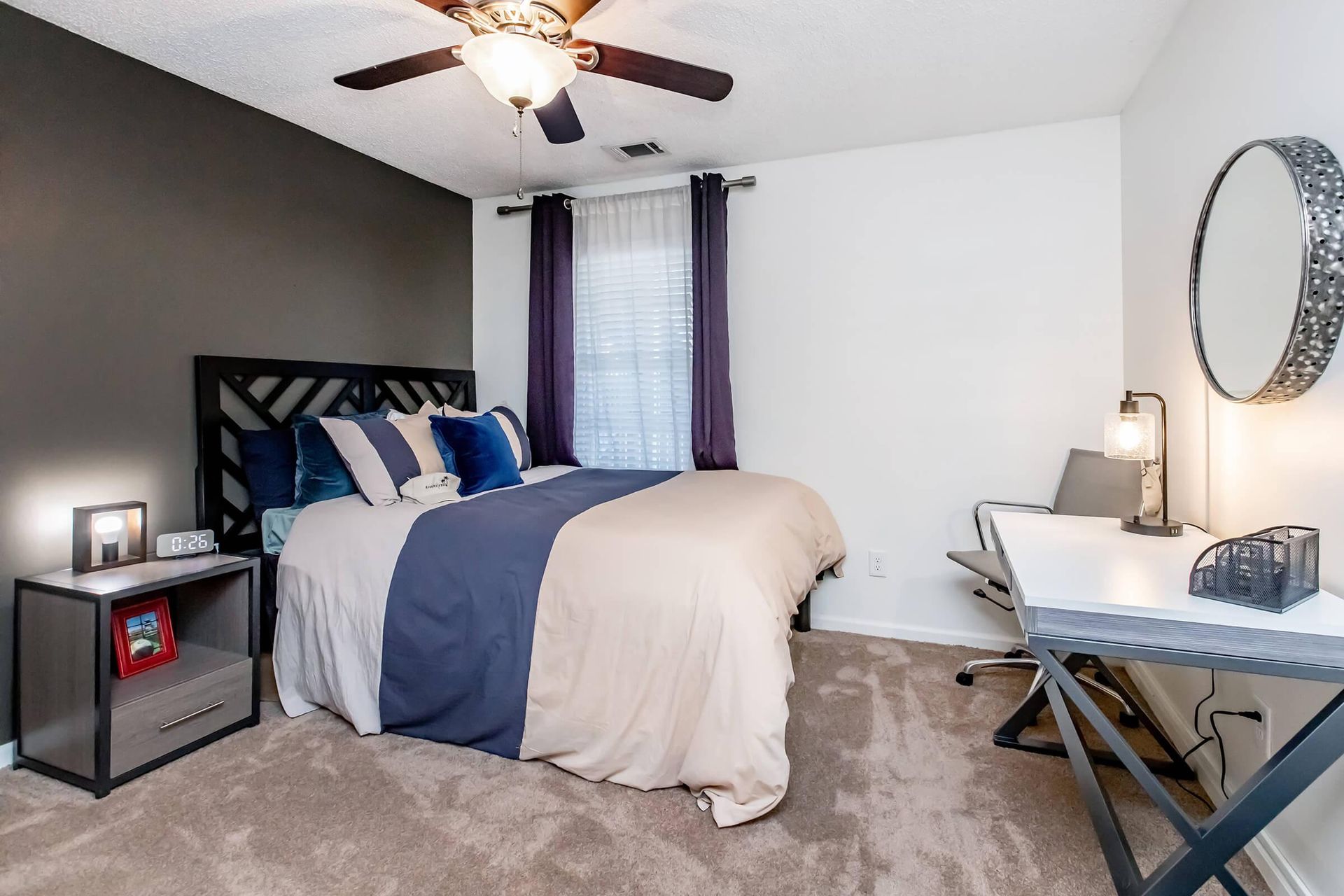 Bedroom with bed, desk, and mirror; dark accent wall, neutral carpet.
