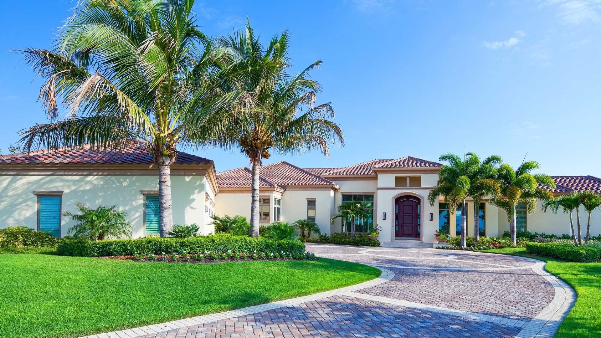 A single-story beige stucco house with a tiled roof, surrounded by palm trees and a landscaped lawn with a stone driveway.