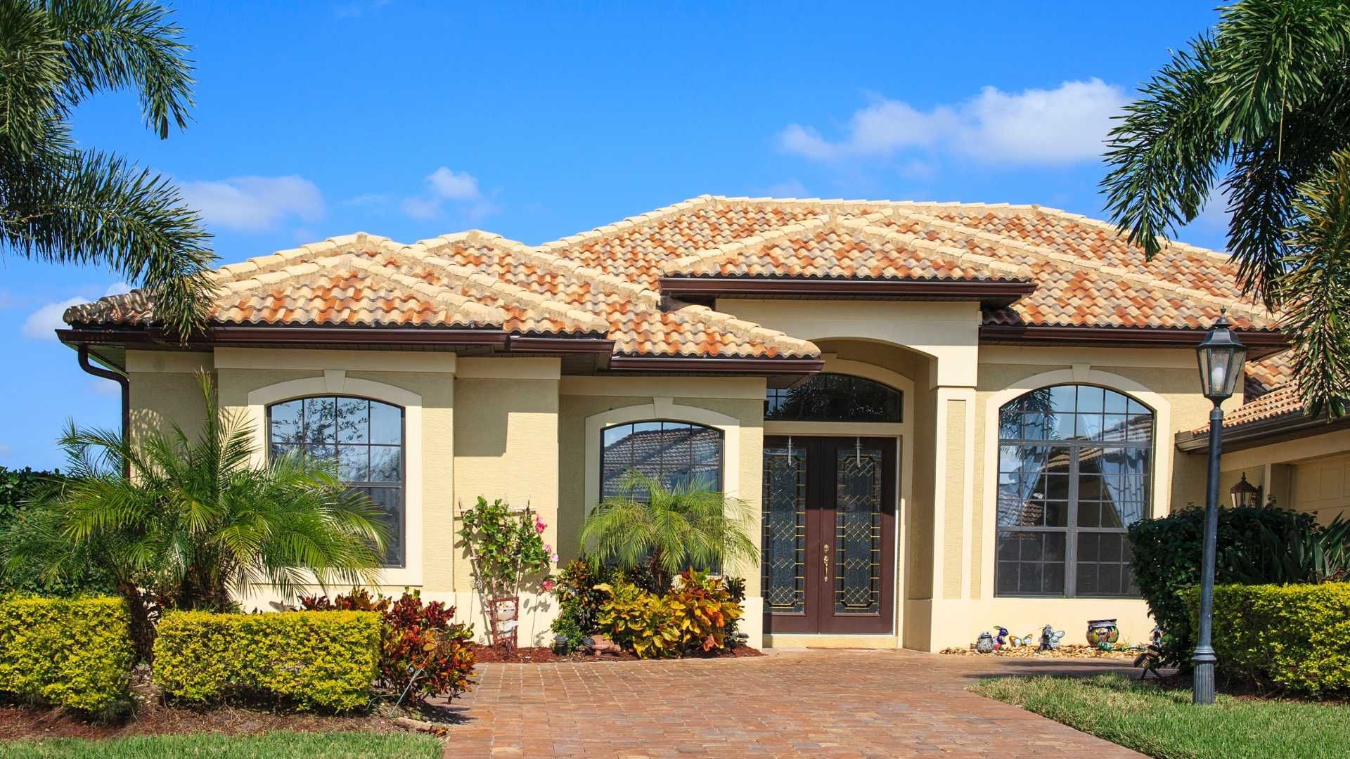 A tan, single-story house with a textured roof, large windows, and a paved walkway, surrounded by lush green landscaping.