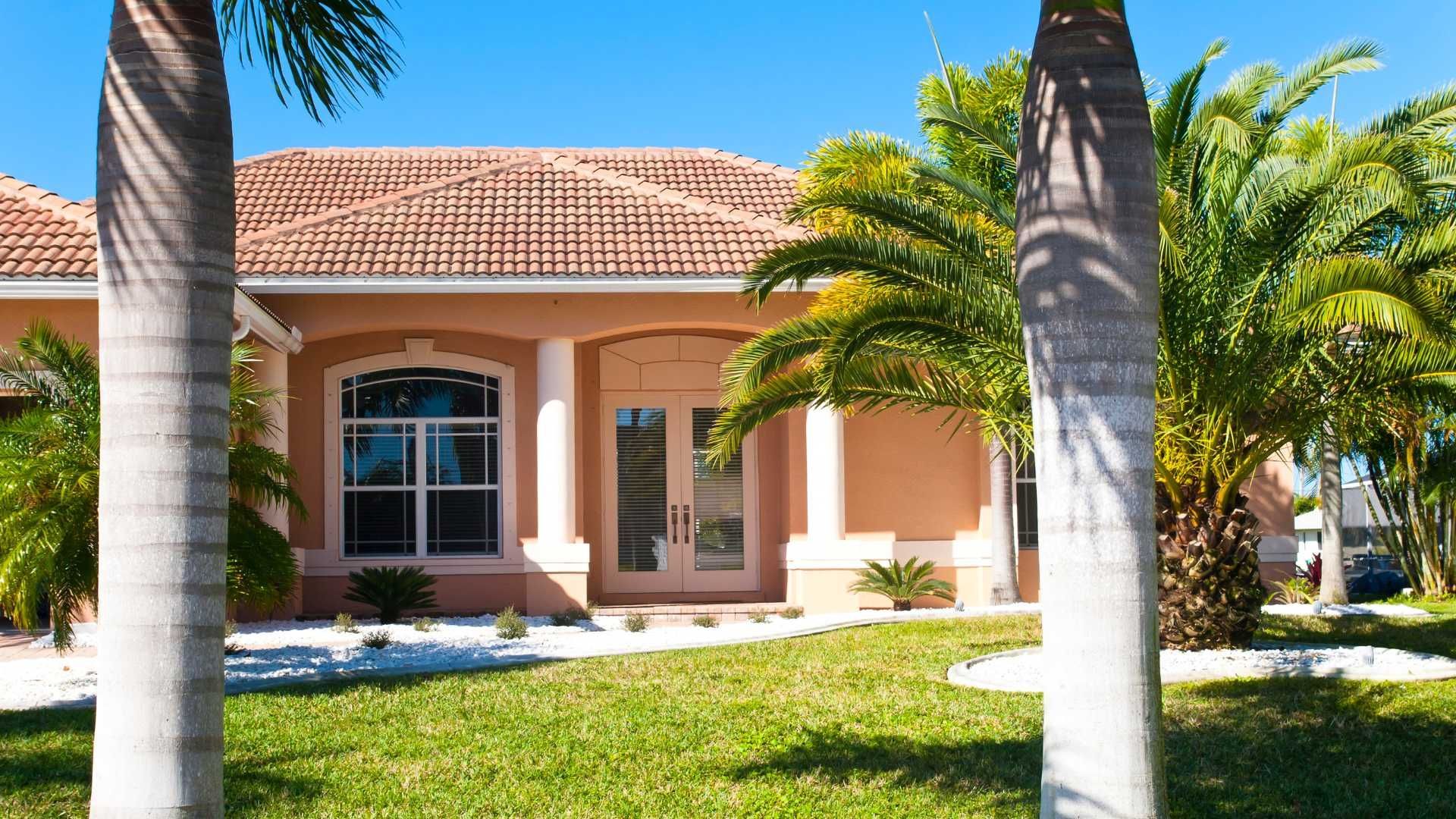 A single-story peach-colored house with a tiled roof, white columns, double doors, and palm trees in the front yard.