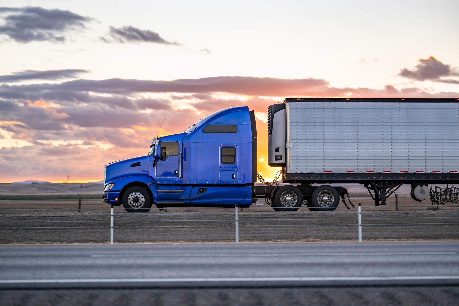 A blue semi truck is driving down a highway at sunset.