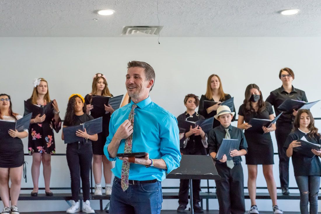 A choir conductor in a blue shirt stands before a group of choir members holding music folders in a rehearsal room.