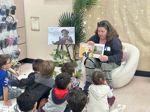 A person reads a book aloud to a group of young children sitting on the floor in a brightly lit indoor classroom.
