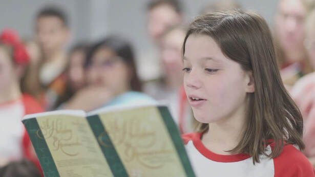 A person in a red and white shirt reads aloud from a green book, with other people blurred in the background.