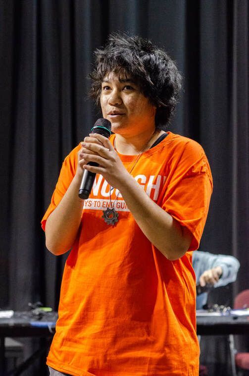 A person with dark, short hair wears an orange shirt and speaks into a microphone against a black curtain background.