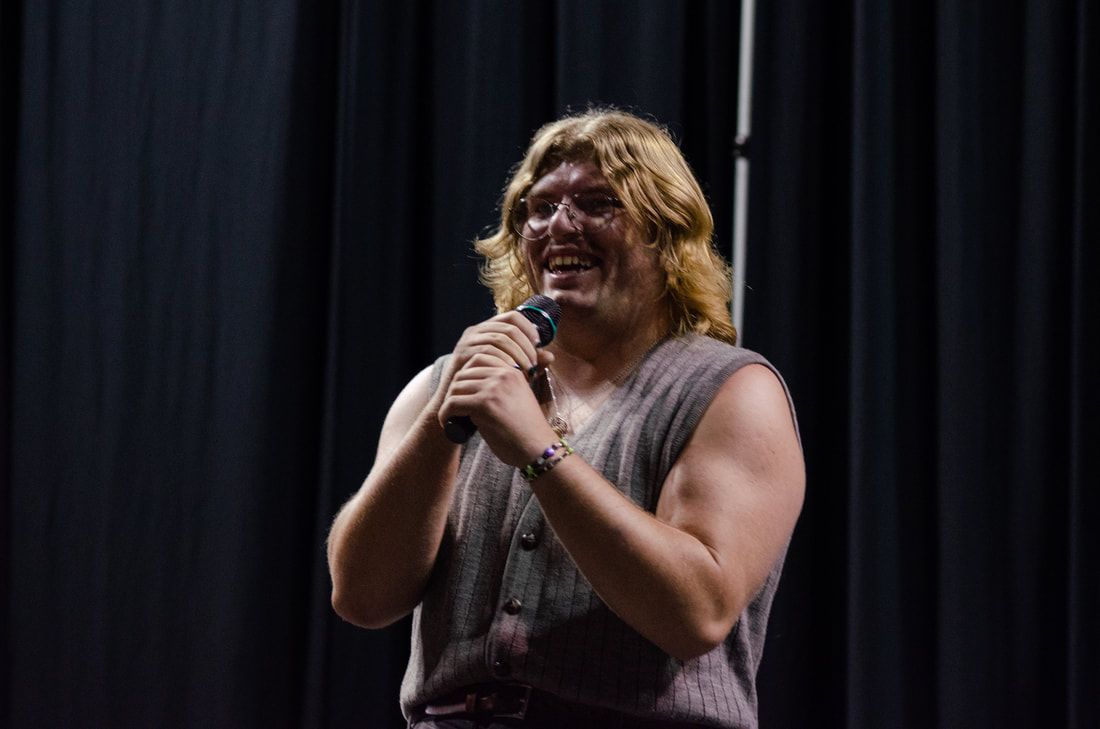 A person with shoulder-length blonde hair, wearing a sleeveless textured top, smiles while holding a microphone onstage.
