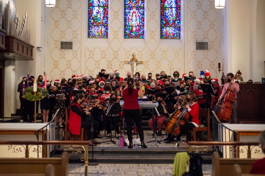 A conductor directs a choir and musicians in a church during a holiday performance before a stained-glass altar.
