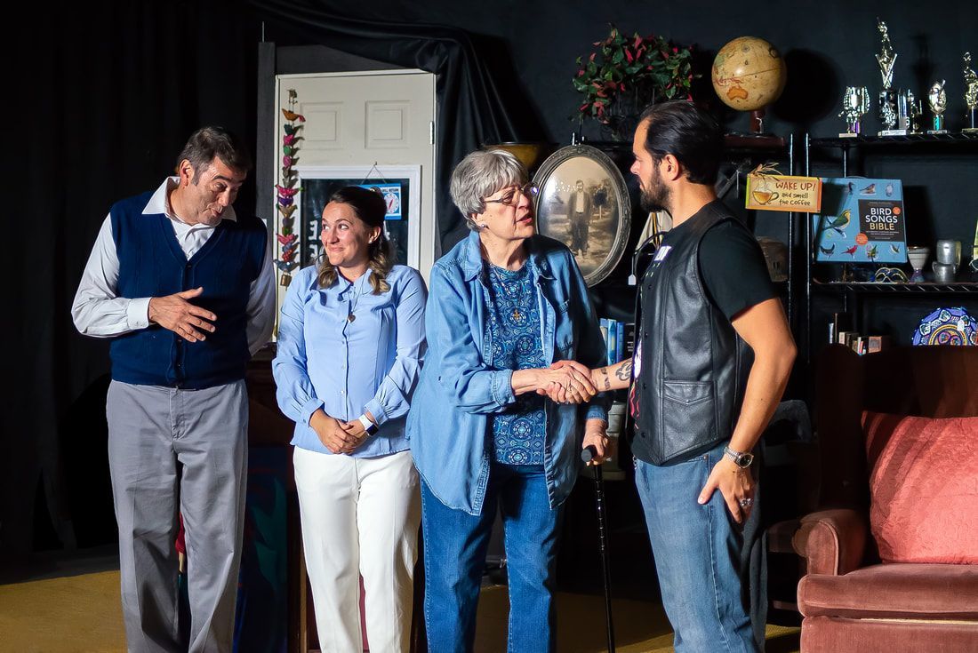 Four individuals stand in a room, with one person shaking hands with another while two others look on.