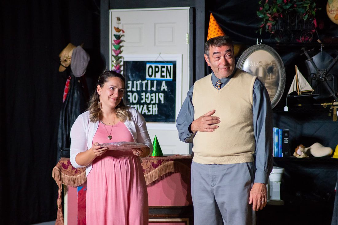 A woman in a pink dress stands next to a man in a sweater vest and party hat in a room decorated like a small shop.