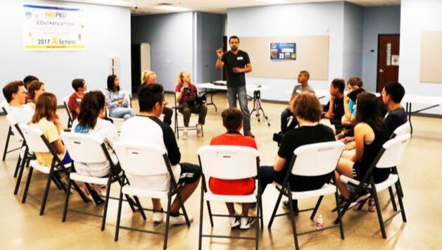 A group sits in a circle in a room, listening as a person stands and gestures to facilitate the discussion.