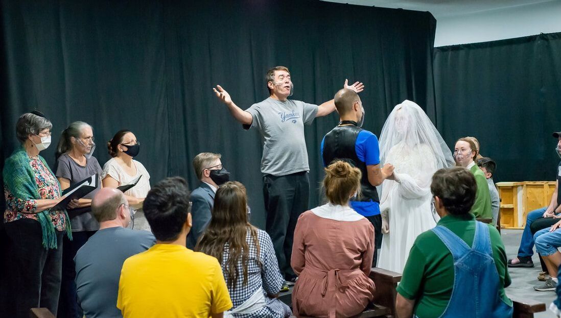 A director gestures while leading a theater rehearsal with a cast on a dark stage, including a person in a wedding veil.