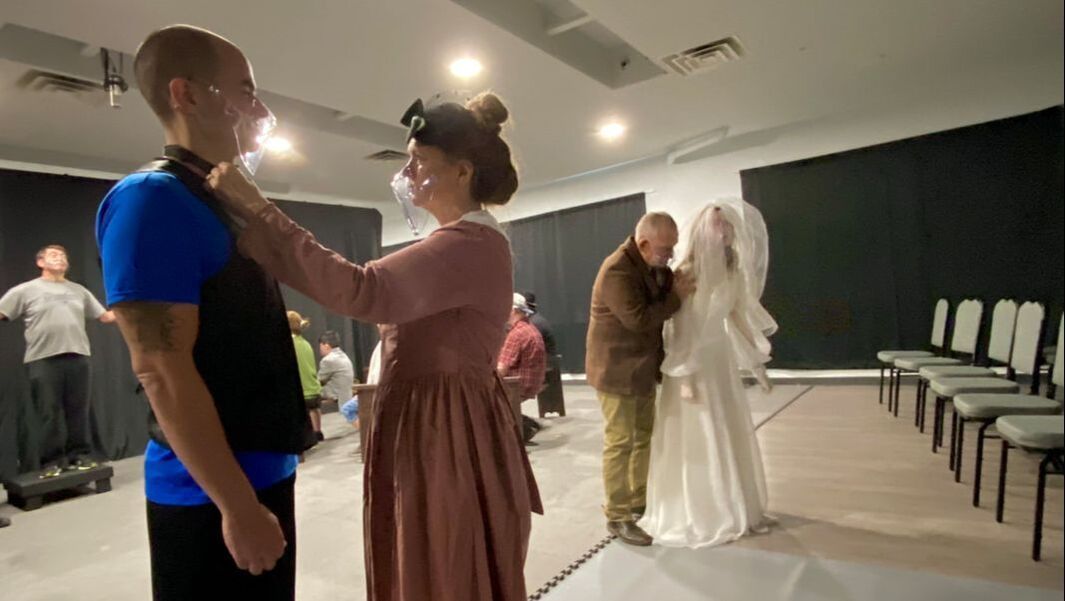 A person in a pink gown adjusts a collar on a man, while another person helps a person in a white wedding dress nearby.
