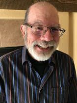 A person with a beard and glasses wearing a striped button-down shirt, smiling warmly indoors.