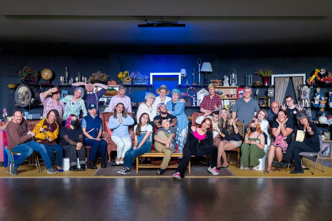 A diverse group of people posing together on a stage filled with theatrical props, shelves of objects, and studio lighting.