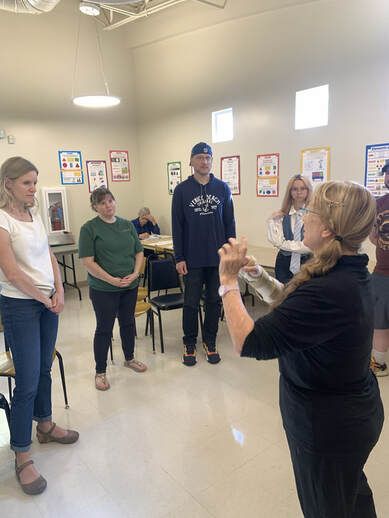 A group of people standing in a circle in a bright room, listening to a speaker in the foreground.
