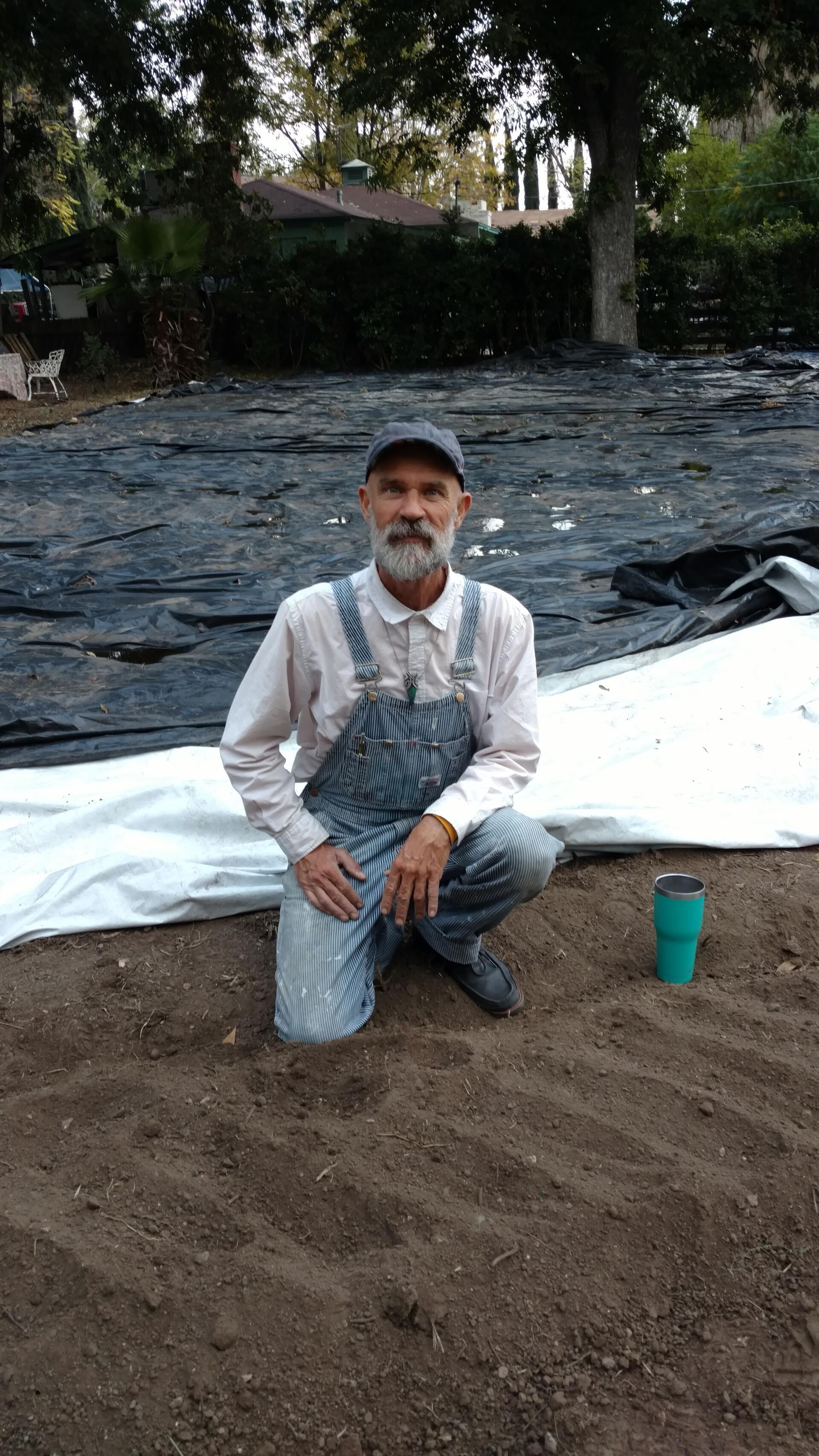 Man in overalls kneels on tilled soil.  He wears a cap and has a beard.  A teal cup sits nearby. Outdoor setting.