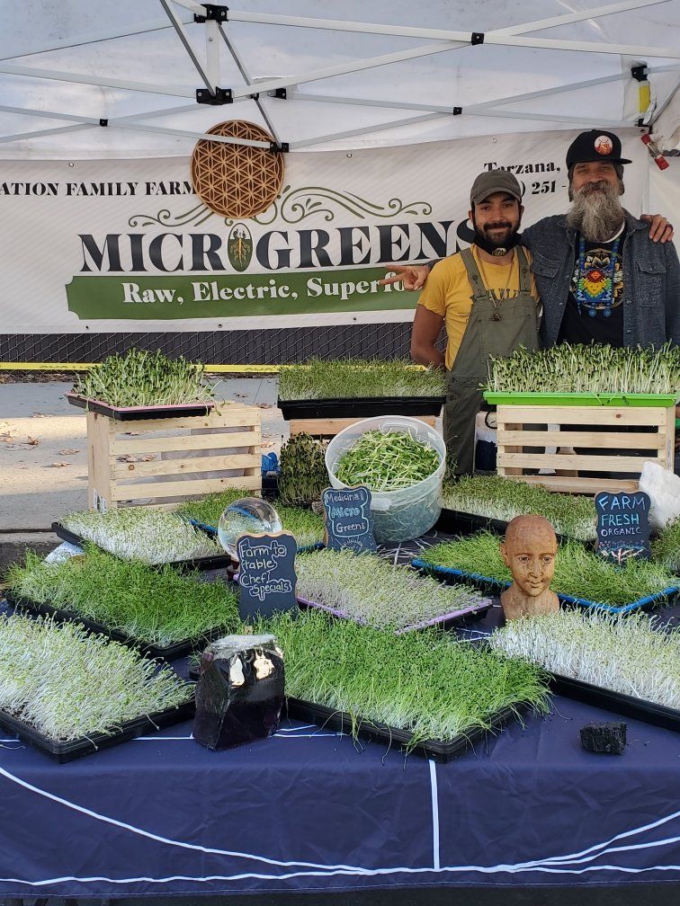 Two men stand at a farmer's market booth selling microgreens. A sign reads 