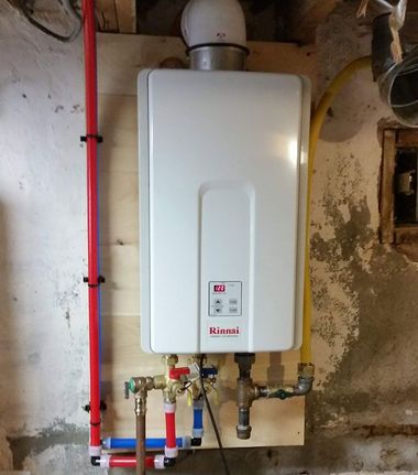 A tankless water heater on a plywood board with red and blue pipes and a vent, in a basement.