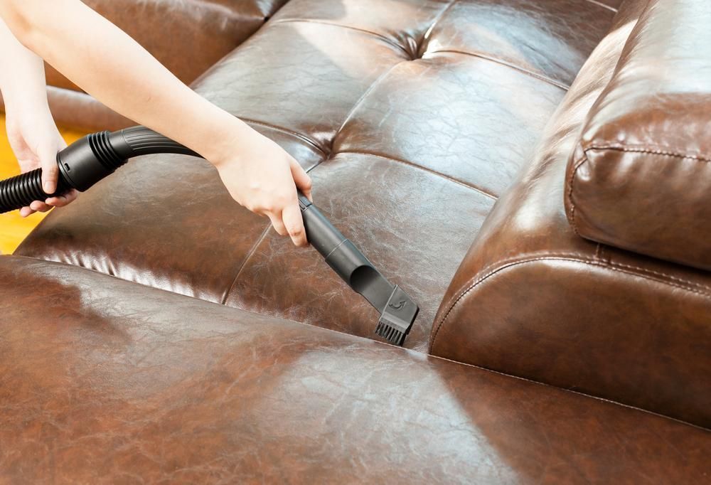 A Person is Cleaning a Brown Leather Couch With a Vacuum Cleaner — Great Lakes Carpet Cleaning & Restoration In Hallidays Point, NSW