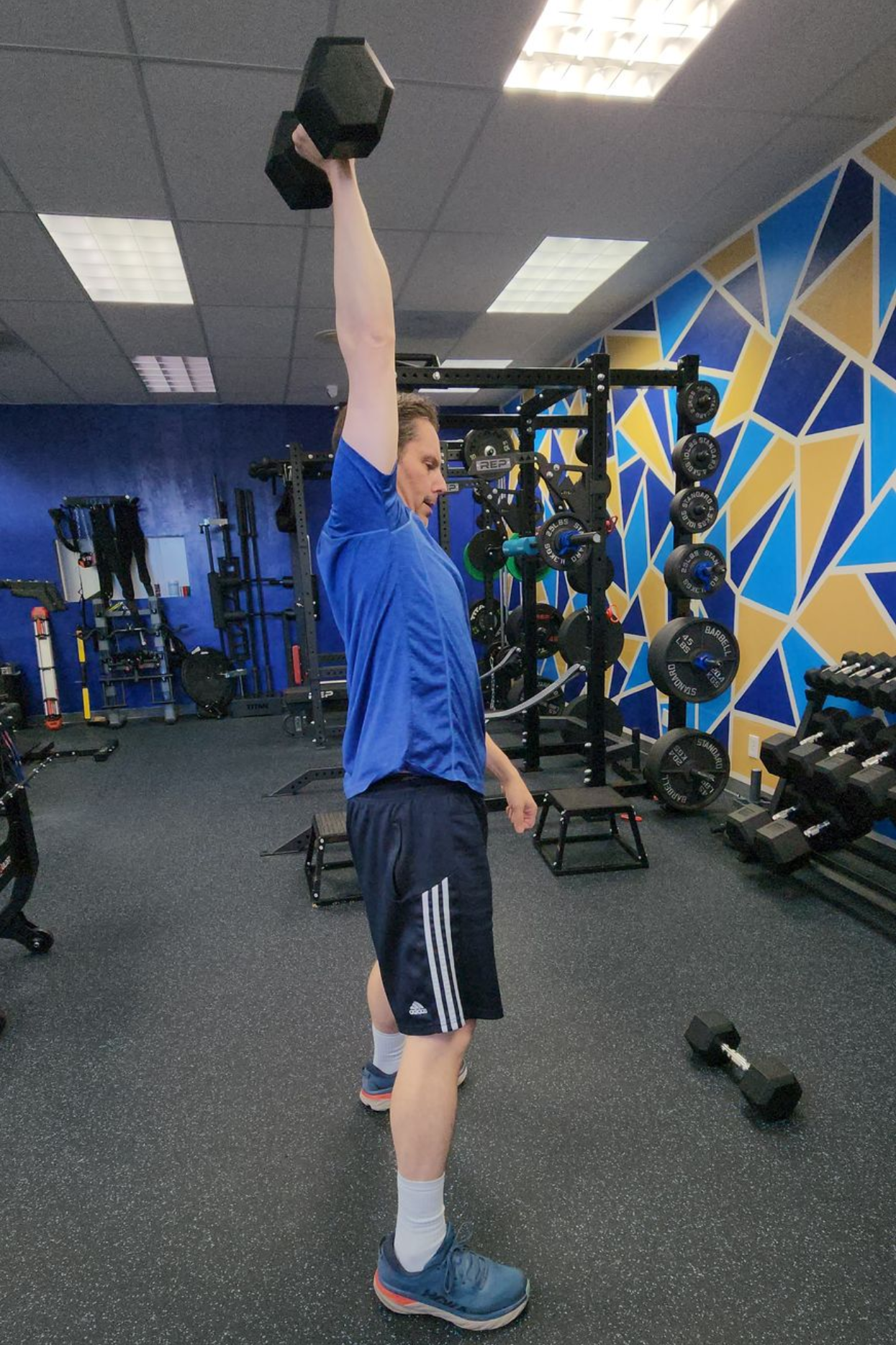 Man performing a single arm dumbbell overhead press in the gym, right arm locked out, strong posture, equipment in the background.