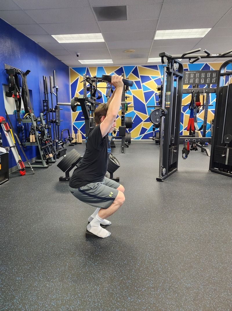 Man performing a squat with a weight overhead on a balance pad to improve balance and strength in a well equipped and well lit gym