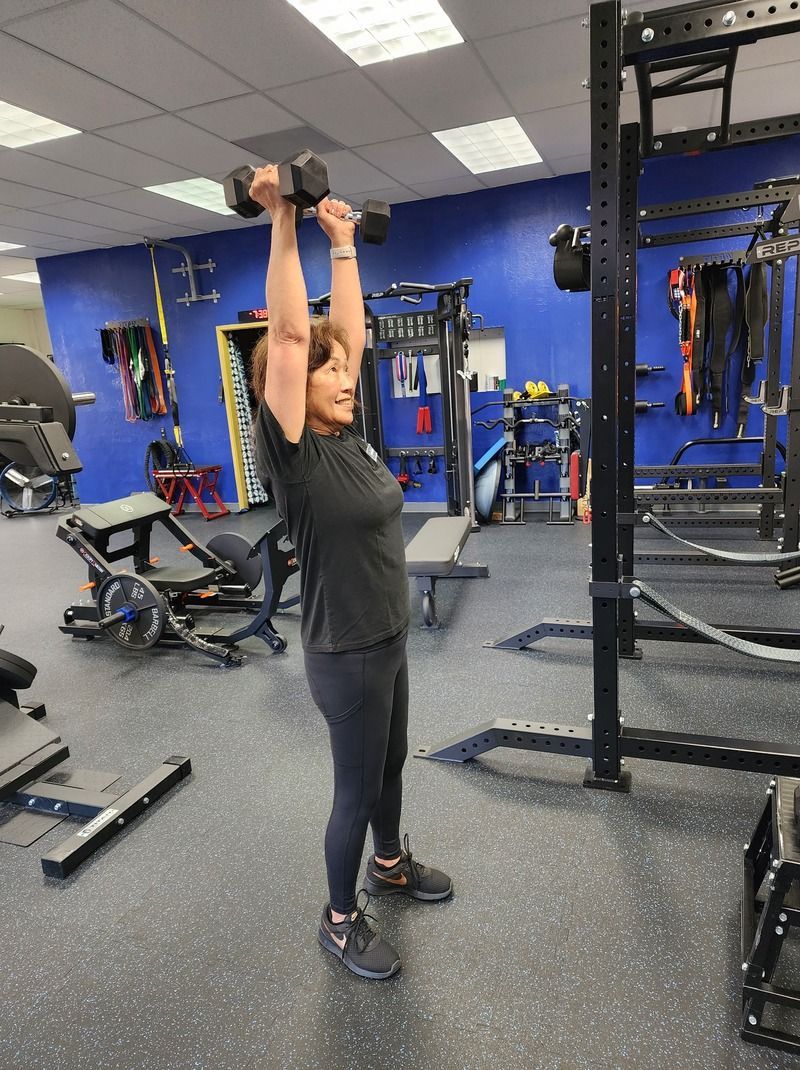 Older lady with a smile on her face wearing gym clothes lifting dumbbells overhead to improve strength and overall well-being in a well-equipped and clean gym.