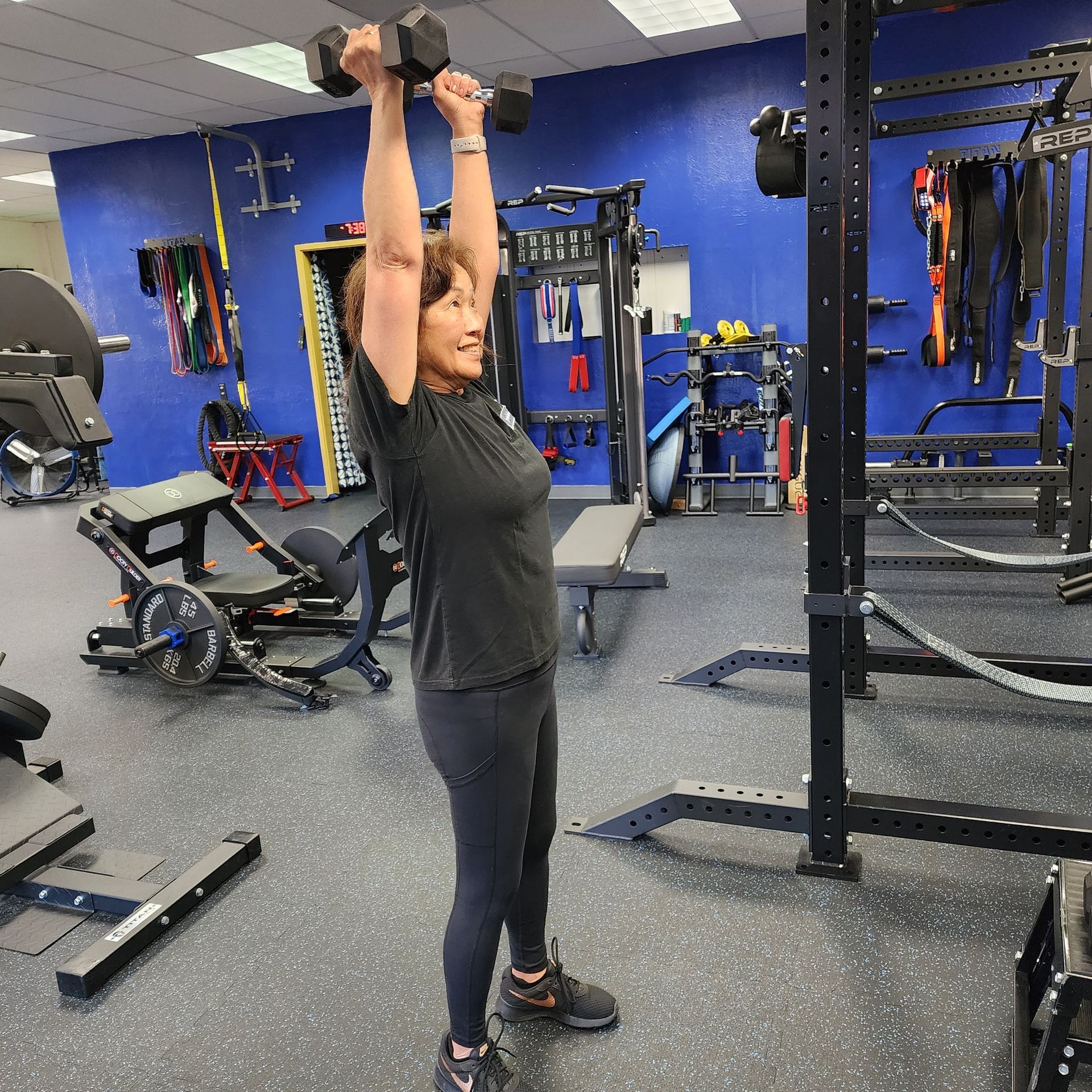 Older lady with a smile on her face wearing gym clothes lifting dumbbells overhead to improve strength and overall well-being in a well-equipped and clean gym.