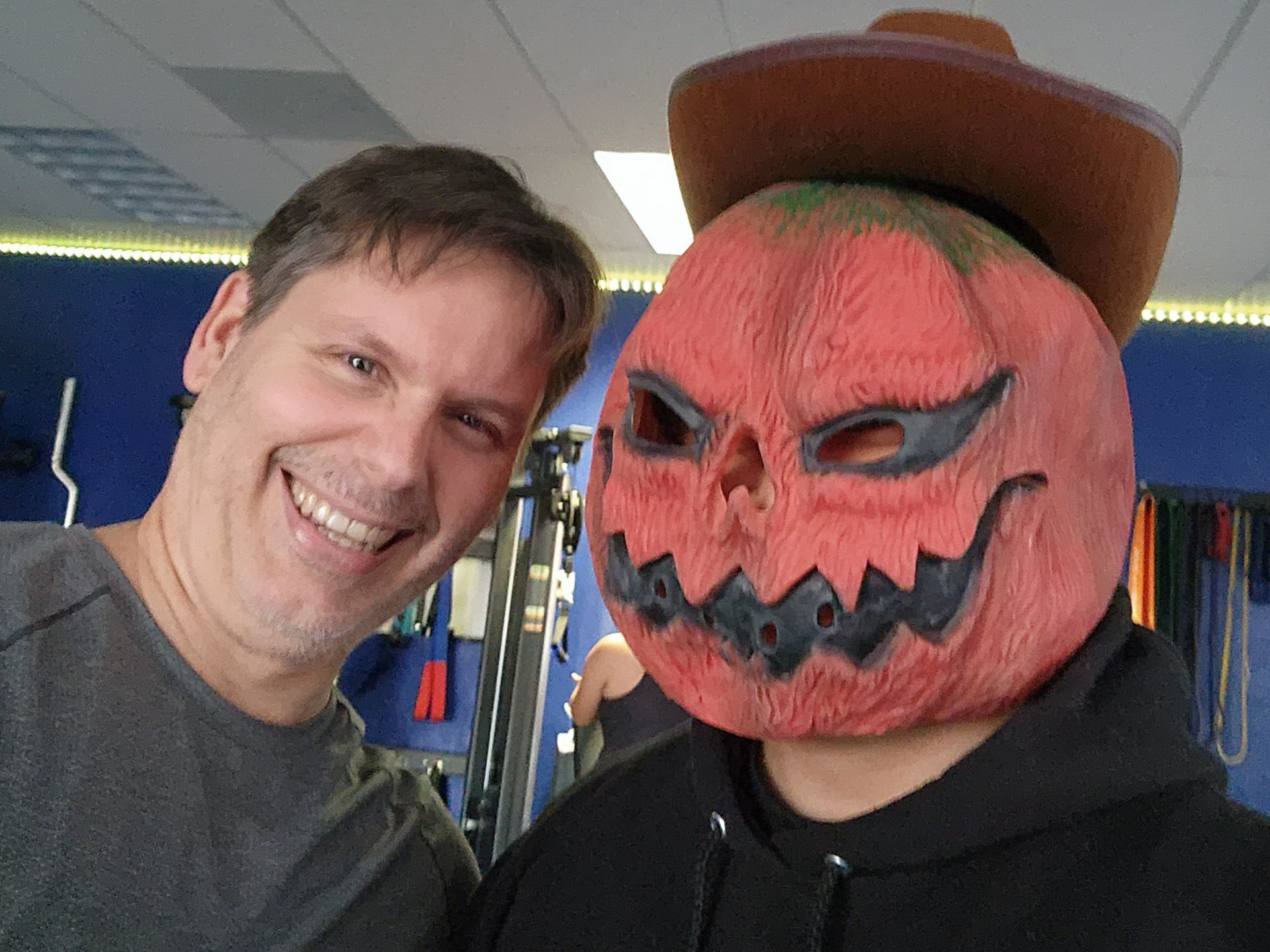 Smiling client next to a coach wearing a pumpkin mask and cowboy hat inside the gym, giving a thumbs up for a fun training moment.