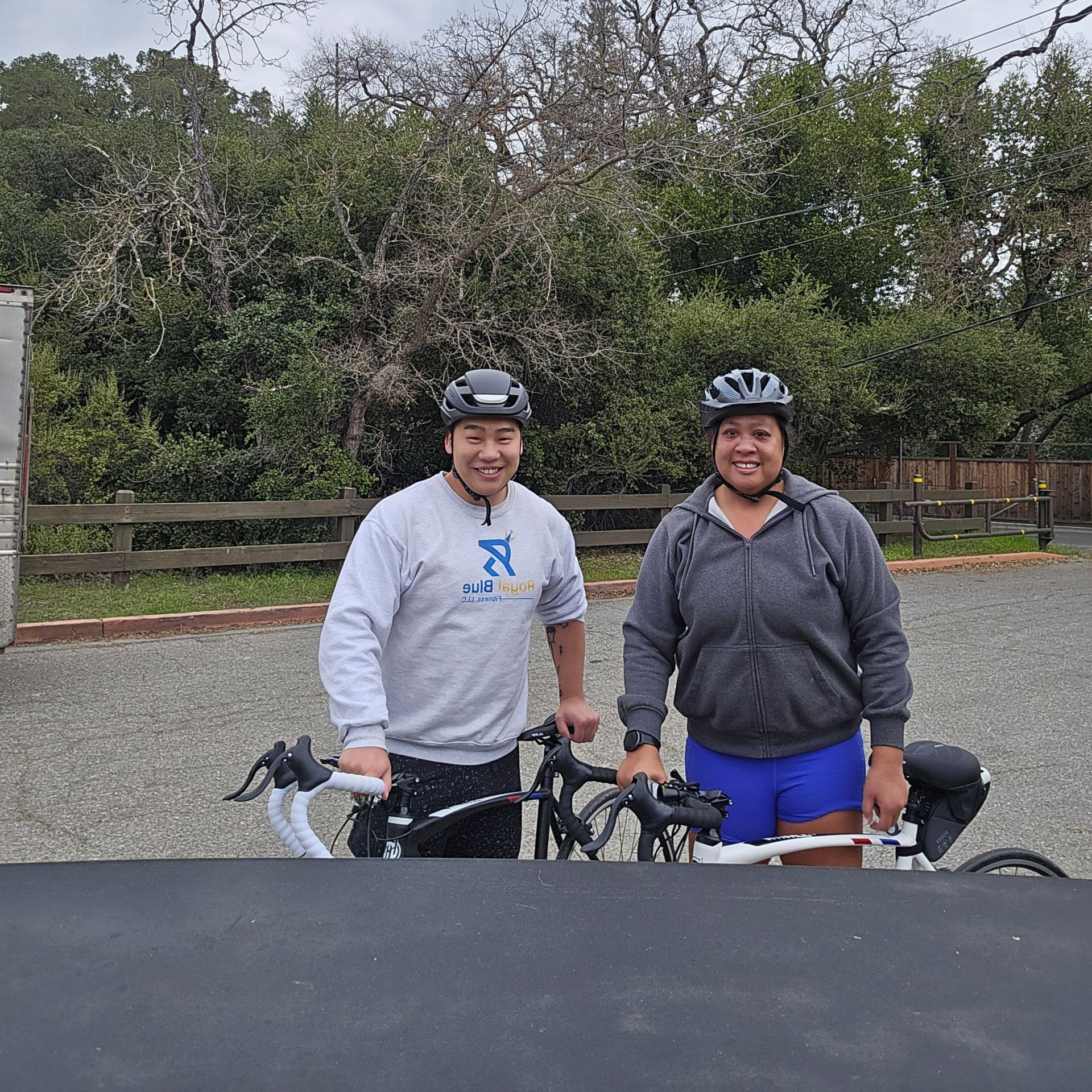 Coach and client Freda with road bikes, wearing helmets and smiling in front of trees during a Royal