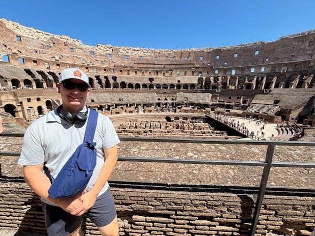 Man wearing sunglasses and a cap at the Colosseum in Rome, standing by a railing with headphones aro