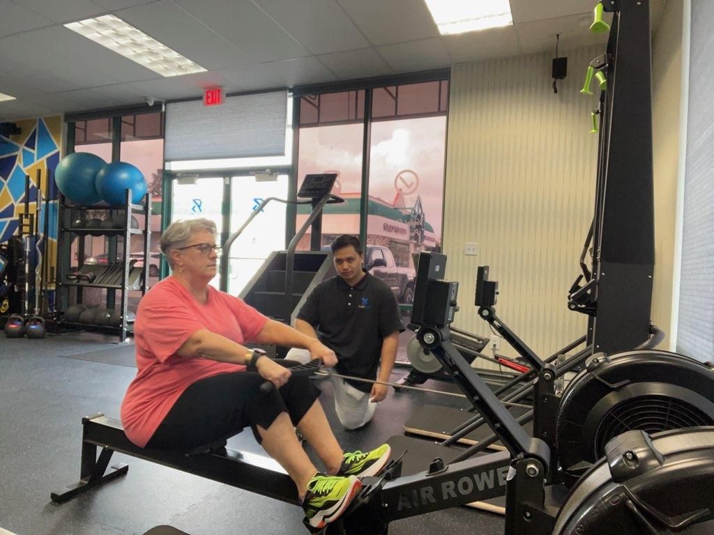 Adult client using a rowing machine while a coach provides form guidance in the Royal Blue Fitness studio.