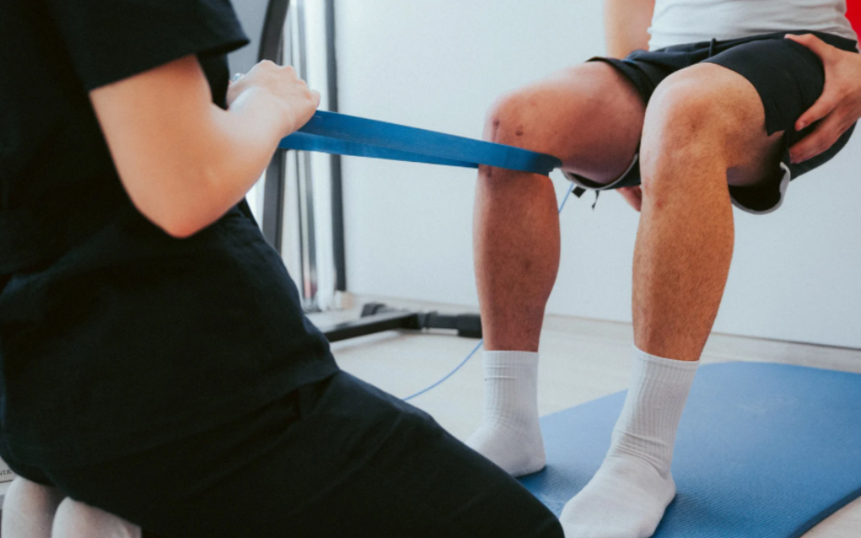 Prehab & post-injury training guided by our structured system. Coach guiding knee rehabilitation with a resistance band as the client performs controlled movement on a blue mat.