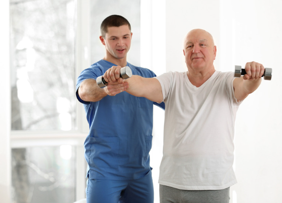 A healthcare professional assists a senior man lifting dumbbells, guiding his arm extension in a bright physiotherapy room.