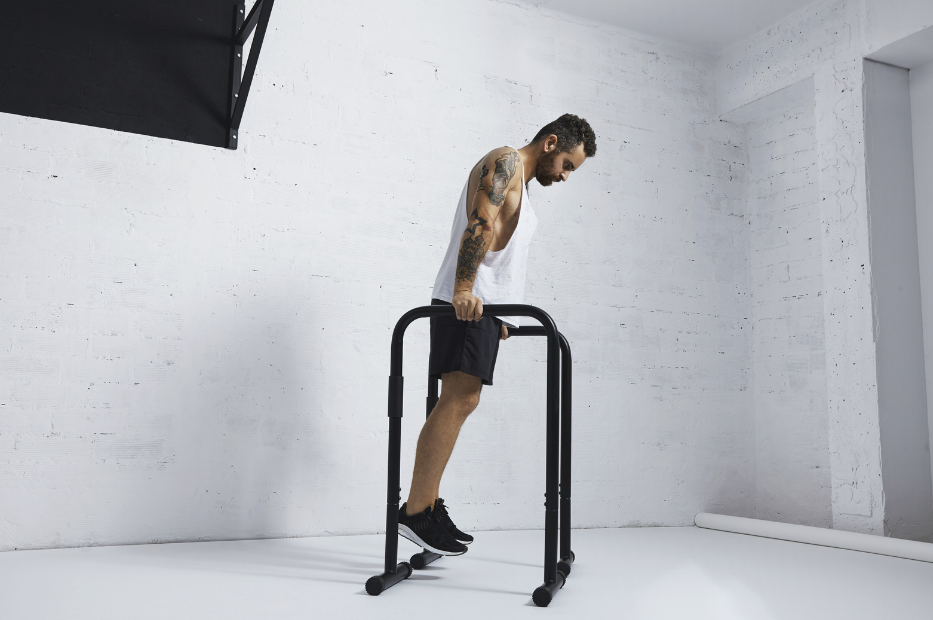 A tattooed man in workout attire leans on a dip bar in a bright, minimalist gym space, preparing for an upper body exercise.