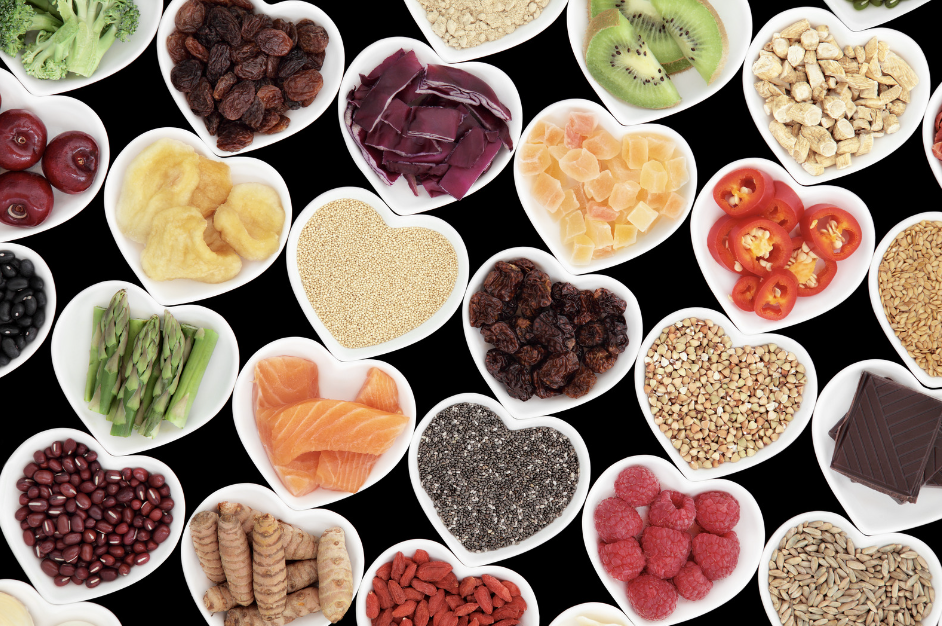 Assortment of healthy foods in heart-shaped dishes symbolizing love for health, including fruits, vegetables, grains, fish, and nuts on a black background