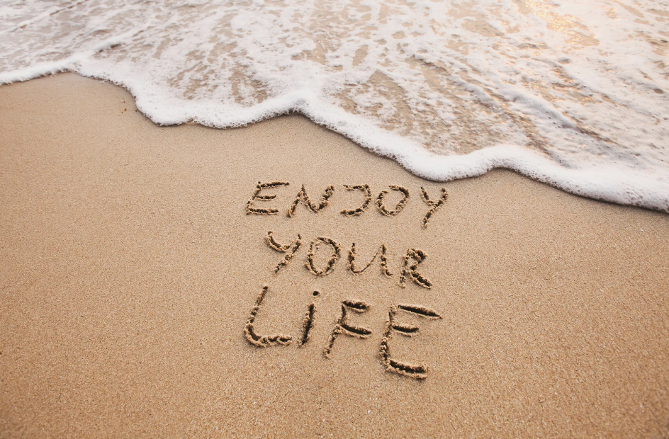 The words 'ENJOY YOUR LIFE' are written in the sand on a beach, with the gentle foam of a wave approaching to wash them away, symbolizing the transient yet beautiful moments of life.