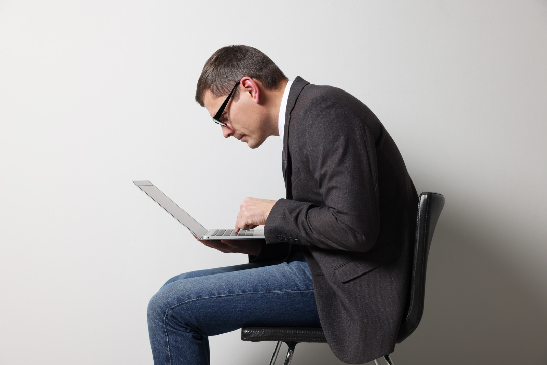 A man in a dark blazer and glasses hunches over a laptop, typing, exhibiting poor posture that could contribute to spinal discomfort or chronic conditions.