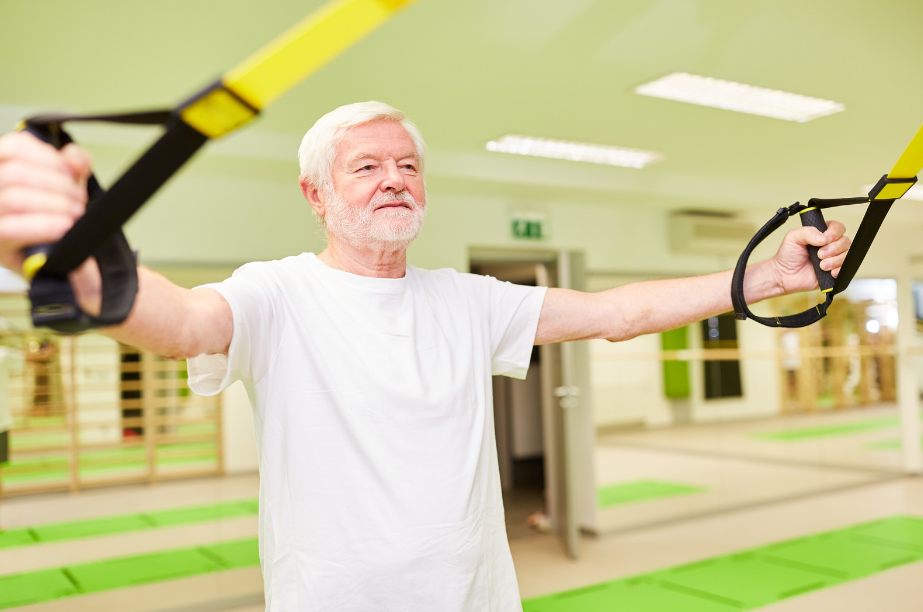 A focused senior man with white hair and a white T-shirt uses suspension straps to exercise in a spacious, well-lit gym, symbolizing a holistic and adaptable approach to maintaining ageless strength and fitness