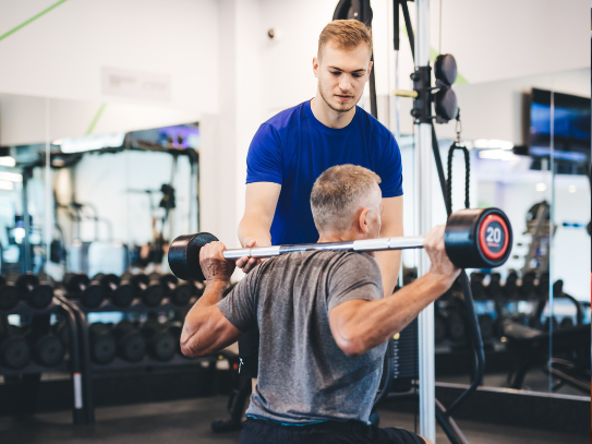 A personal trainer in a blue shirt attentively guiding a senior client through a barbell exercise with proper form for safety