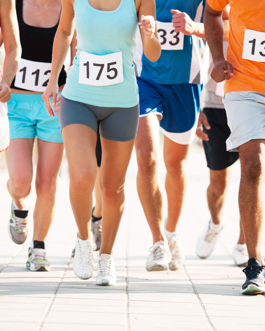 Group of runners wearing race bibs jogging together on a sunny day, shown from the waist down.