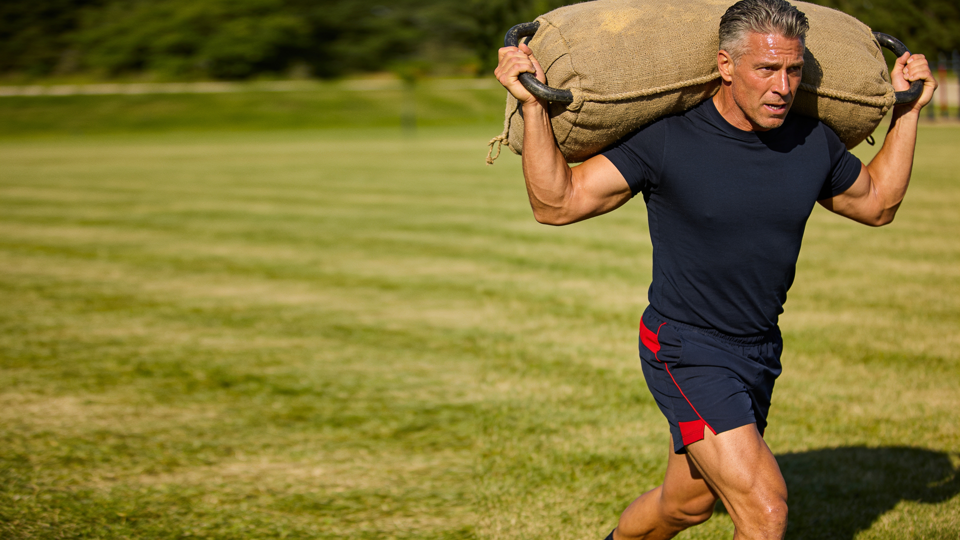 An athletic man carries a heavy training sandbag in each hand across an outdoor field, walking tall 
